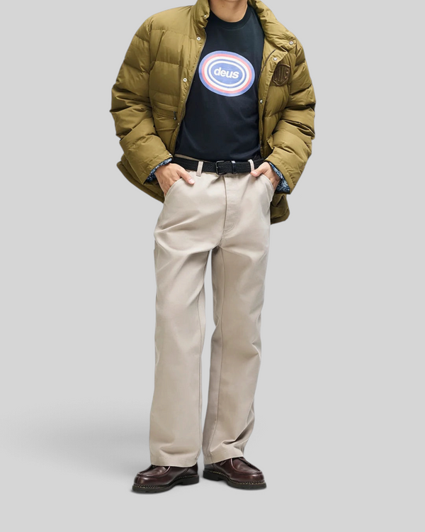 Baffle Haus Carson down jacket in olive with a relaxed fit, worn over a graphic t-shirt and beige trousers.