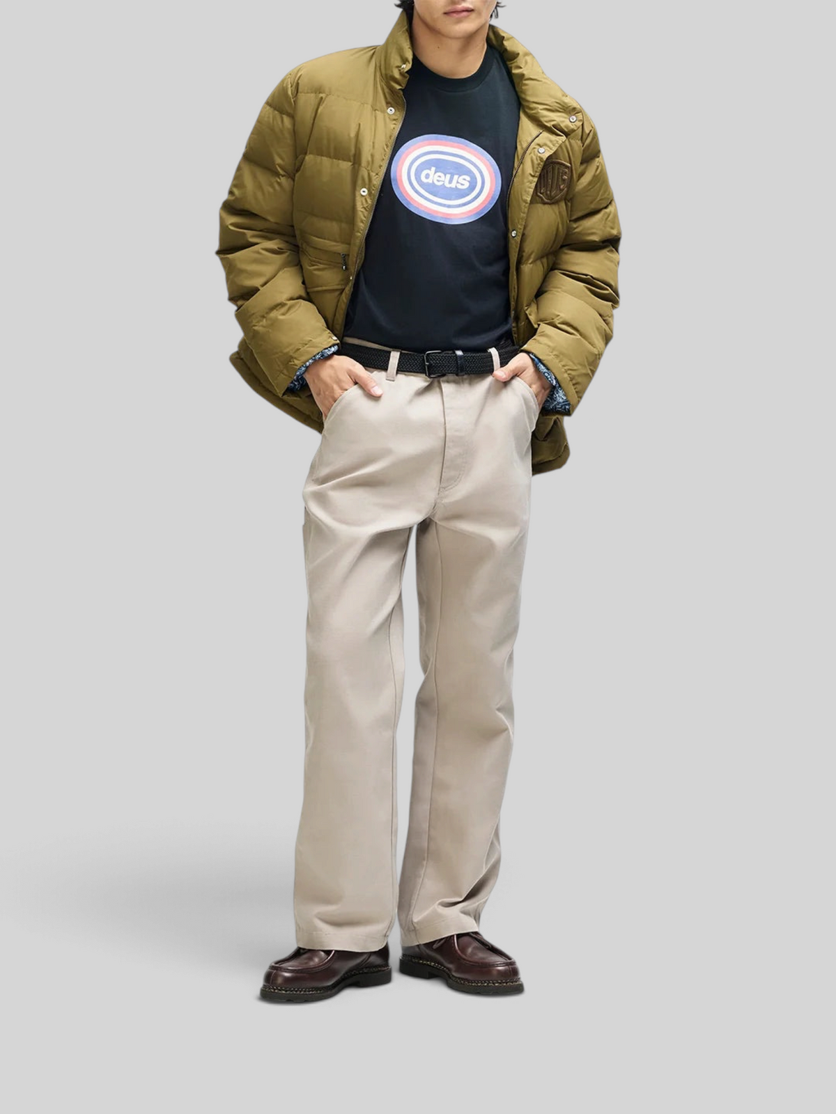 Baffle Haus Carson down jacket in olive with a relaxed fit, worn over a graphic t-shirt and beige trousers.