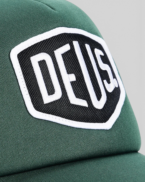 Baffle Haus Baylands trucker cap in racing green with a white and black embroidered Deus logo patch on the front