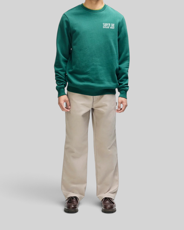 Baffle Haus Chessboxin Crew sweatshirt in racing green with chest graphic, worn with beige trousers.