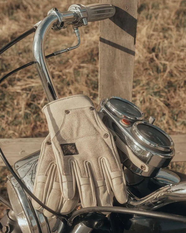 Baffle Haus Age of Glory Crafter leather motorcycle gloves in aged white resting on vintage motorcycle handlebars.