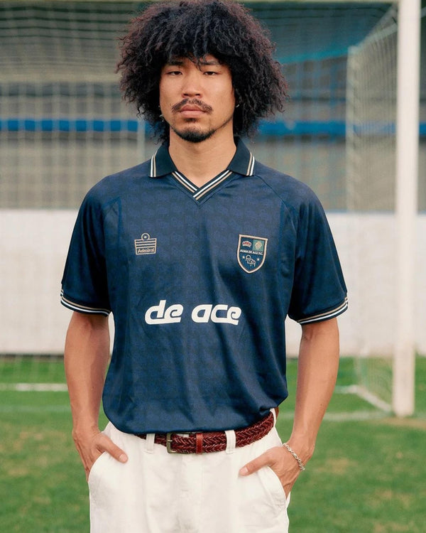 Baffle Haus model wearing a navy blue Alma De Ace F.C. home football shirt with a collared neckline on a pitch.