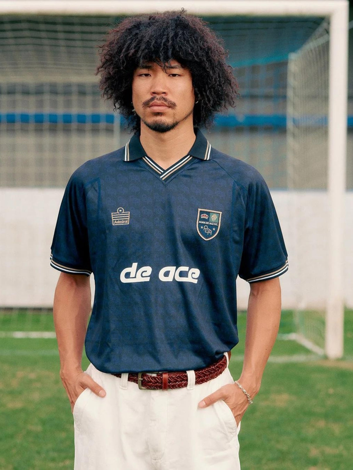 Baffle Haus model wearing a navy blue Alma De Ace F.C. home football shirt with a collared neckline on a pitch.