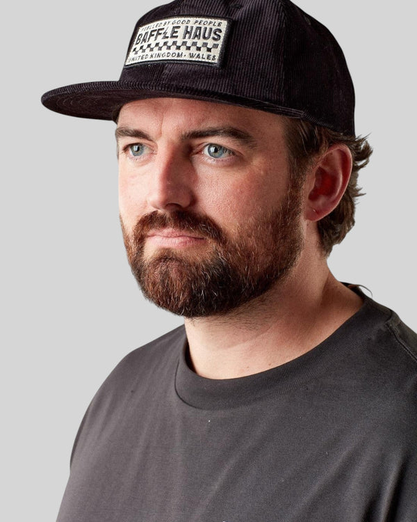 Man wearing a black Baffle Haus corduroy panel cap with a branded patch on the front against a grey background.