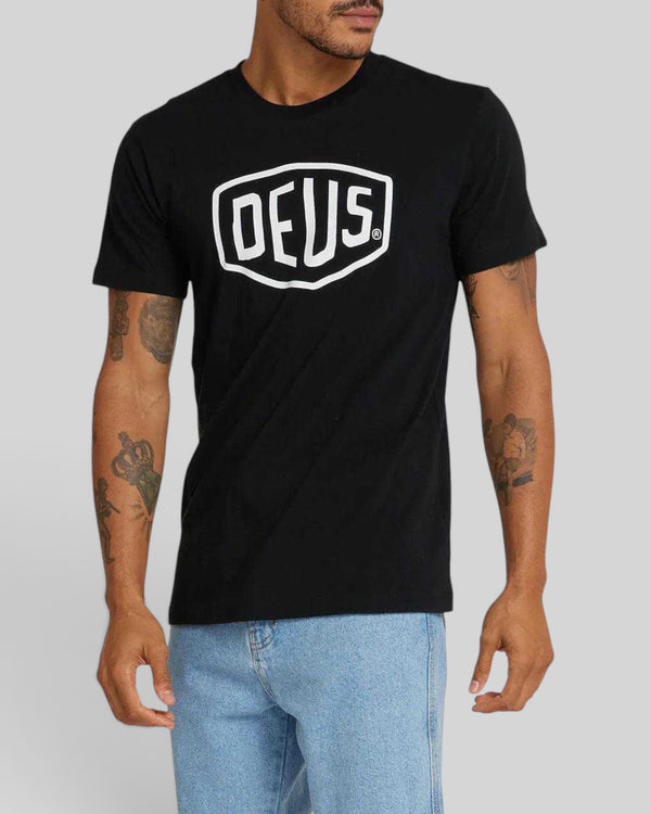 Baffle Haus Shield Tee in black cotton jersey with white Deus shield logo graphic worn by a man in light blue jeans.