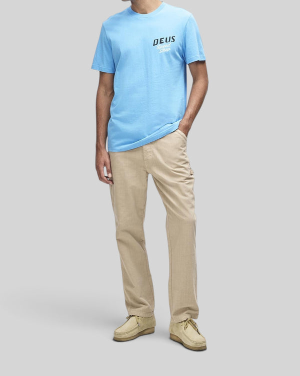 Baffle Haus Dogleg Tee in Bonnie Blue with chest graphic, worn by a model with beige trousers and suede shoes.