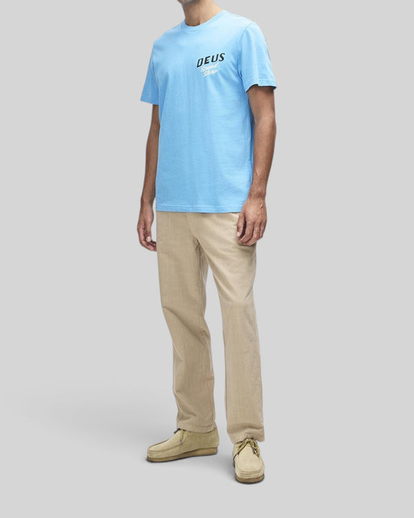 Baffle Haus Dogleg Tee in bonnie blue featuring a chest graphic, worn with beige trousers and suede shoes.