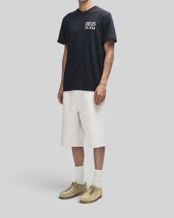 Baffle Haus Camber Tee in black featuring a white chest print, worn with white shorts and beige suede shoes.