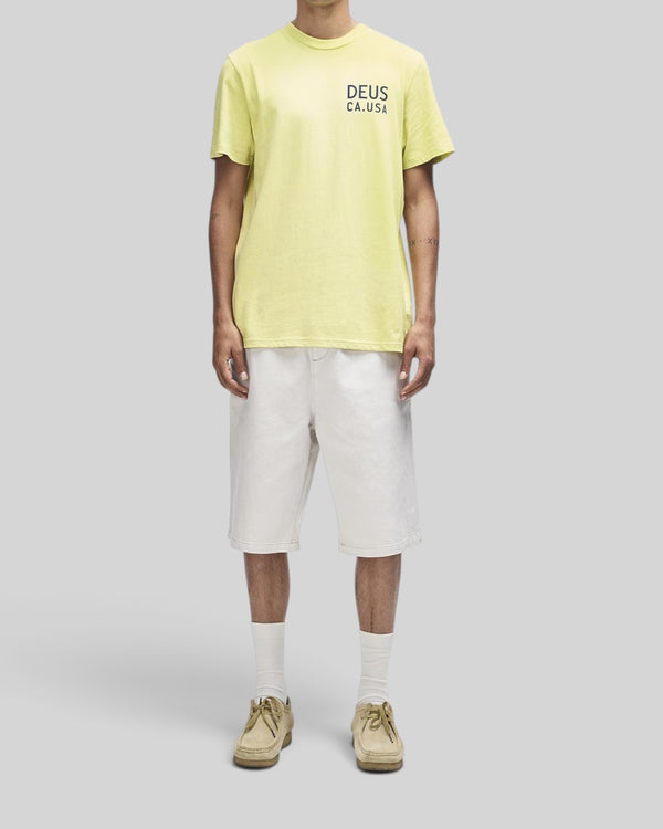 Baffle Haus Camber Tee in canary yellow with chest print, worn with white shorts and tan shoes on a model.