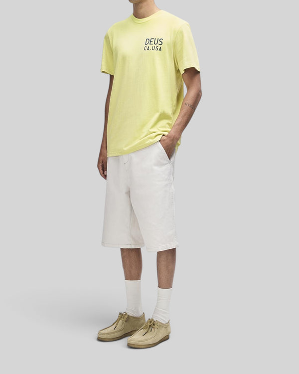 Baffle Haus Camber Tee in canary yellow featuring a chest graphic, worn with white shorts and tan suede shoes.