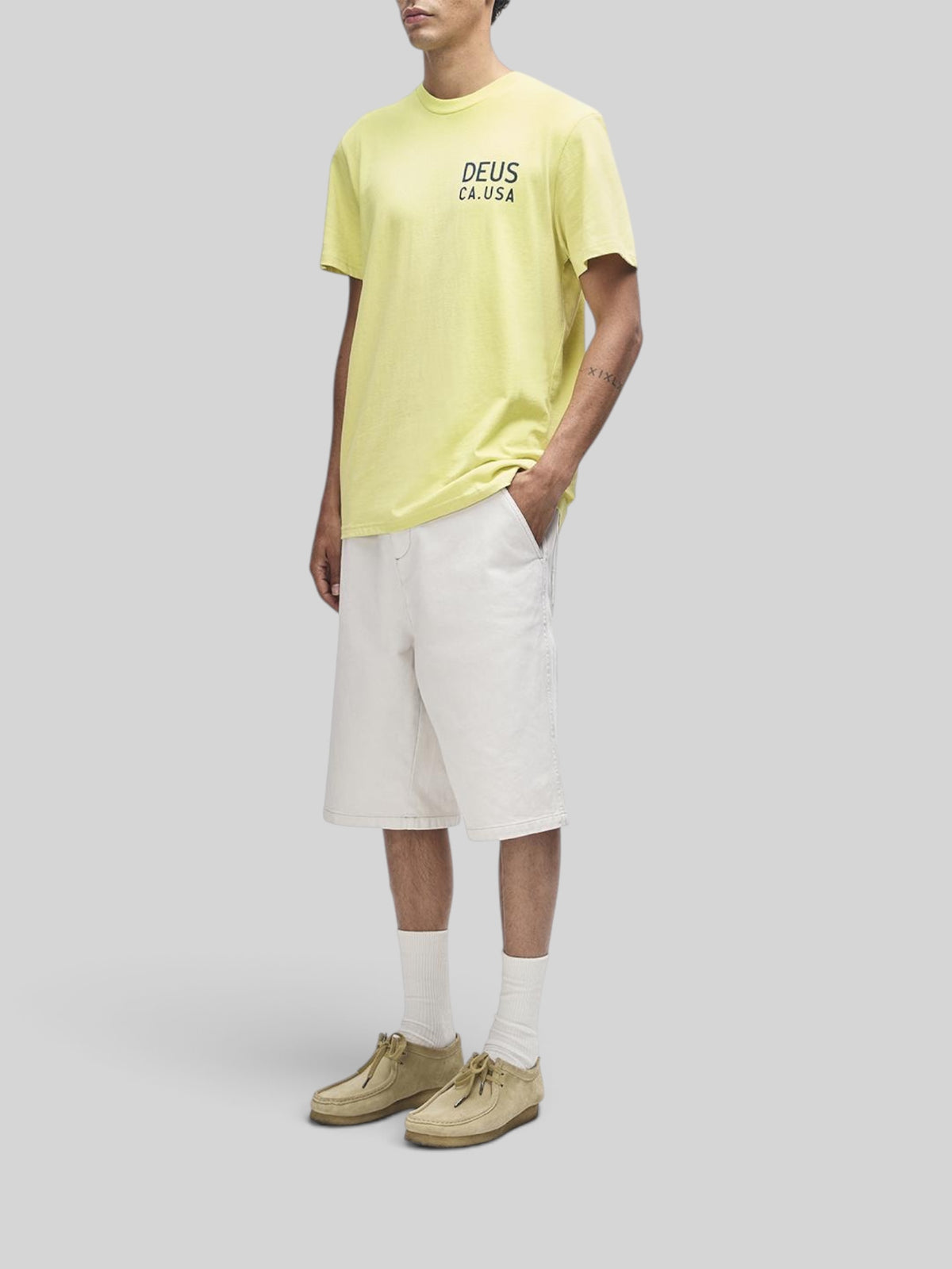 Baffle Haus Camber Tee in canary yellow featuring a chest graphic, worn with white shorts and tan suede shoes.