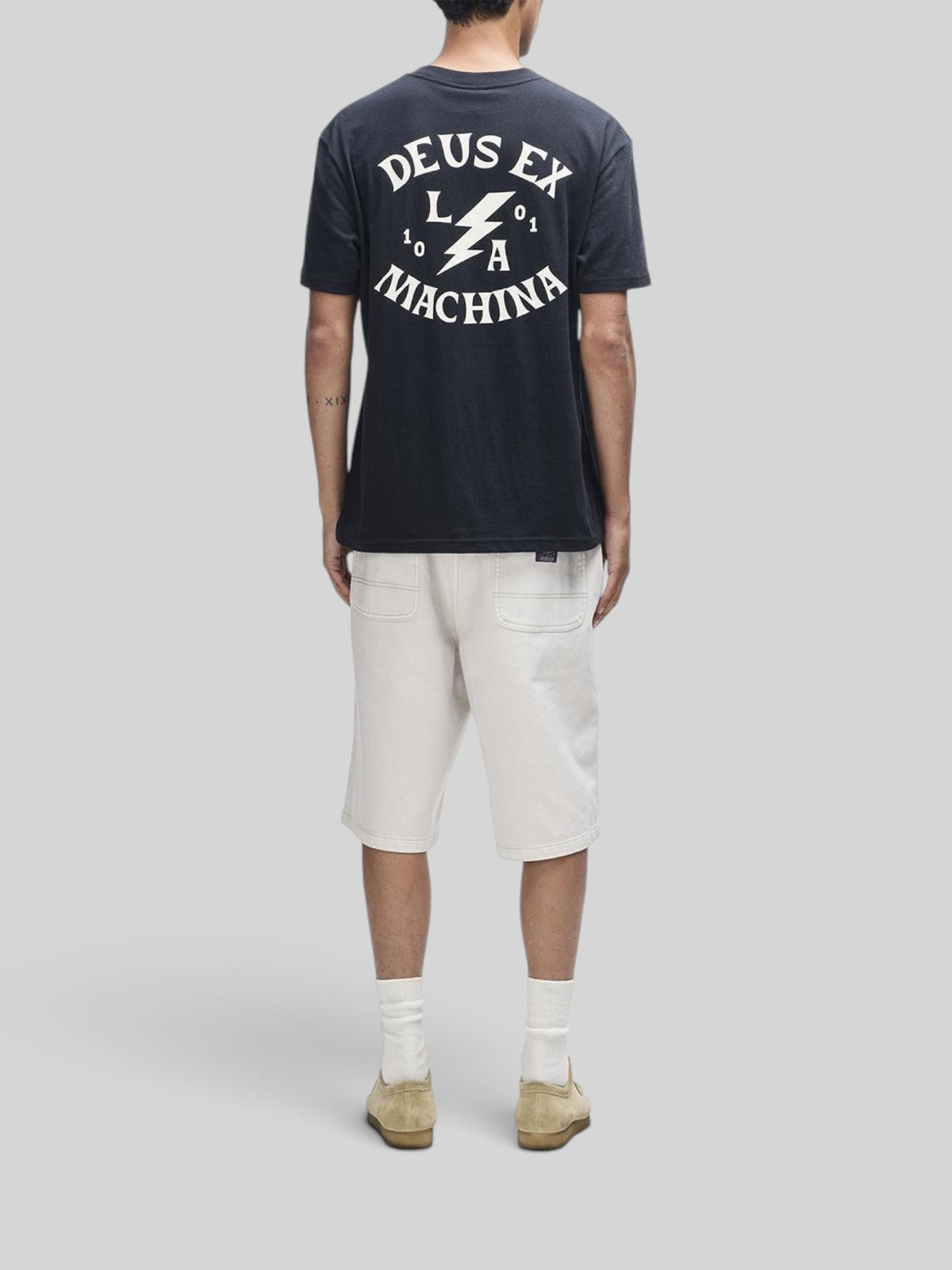 Baffle Haus Chinook black box fit t-shirt with white Deus Ex Machina graphic print on back, worn with cream shorts.