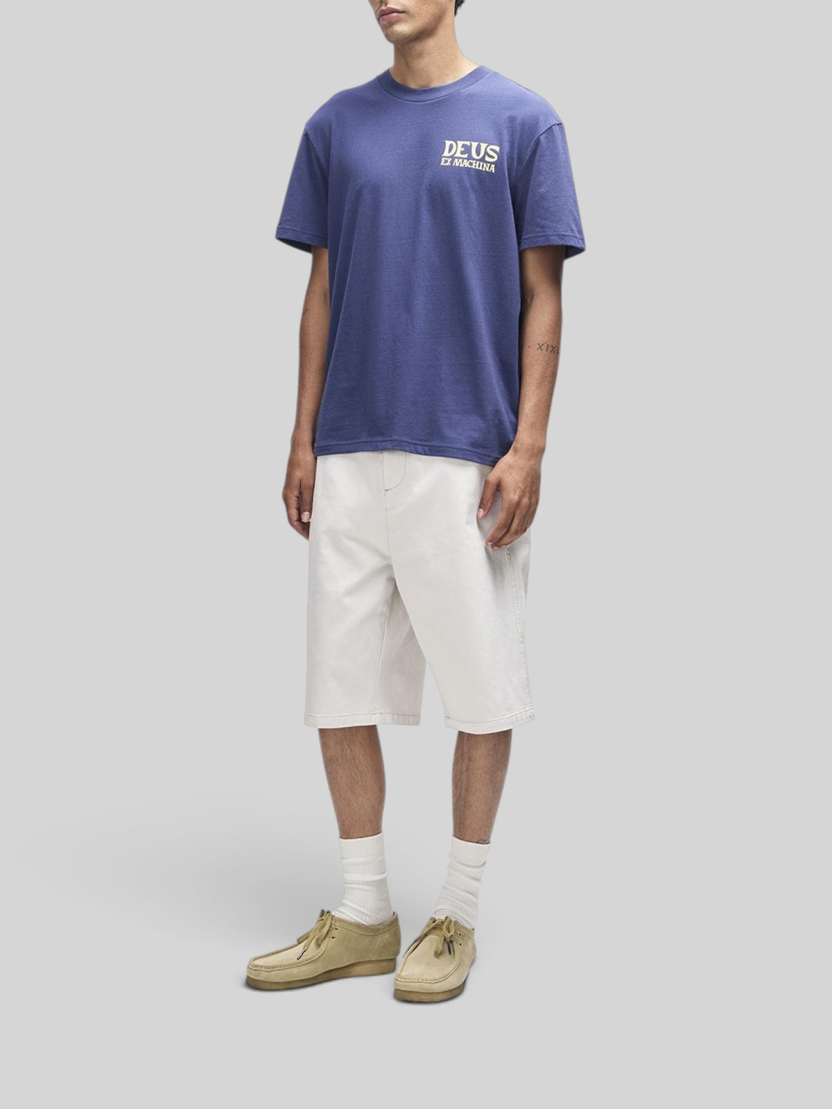 Baffle Haus Chinook Tee in mechanic blue with Deus Ex Machina chest print, worn with white shorts and beige shoes.