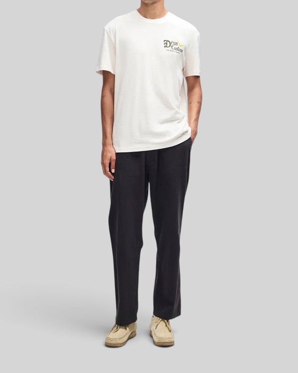 Baffle Haus Beep-Beep Tee in dirty white with chest graphic, worn by a model with black trousers and beige shoes.
