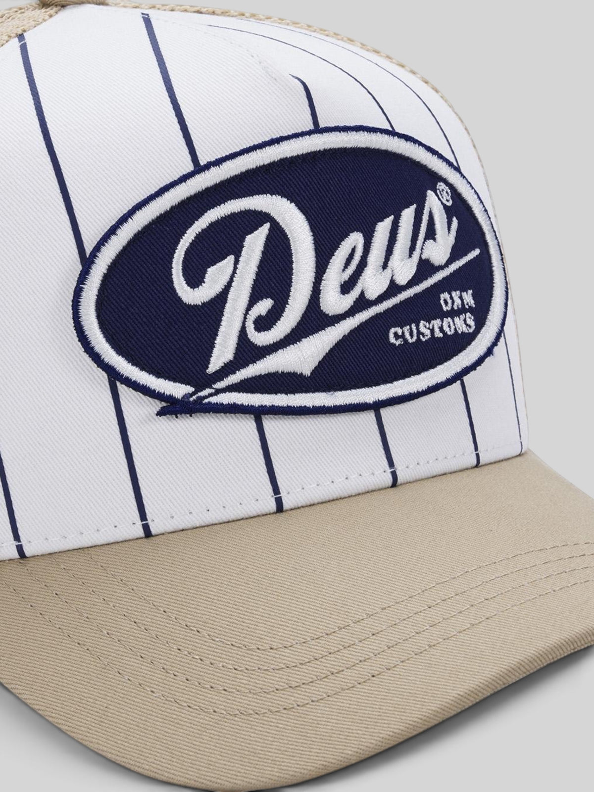 Baffle Haus Forecourt Trucker cap in white with navy pinstripes, tan brim, and Deus Customs oval patch.