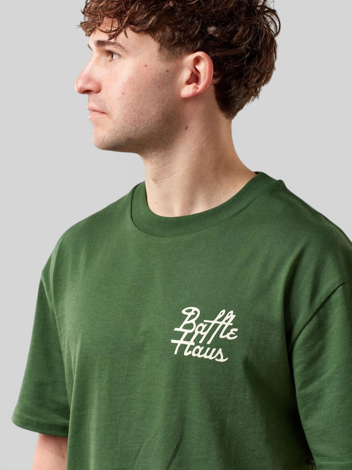Baffle Haus forest green cotton t-shirt with white cursive logo on chest worn by a man in profile view