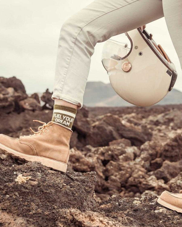 Baffle Haus Fuel Your Dreams socks in olive green with white stripes and text, worn with tan boots and a helmet.