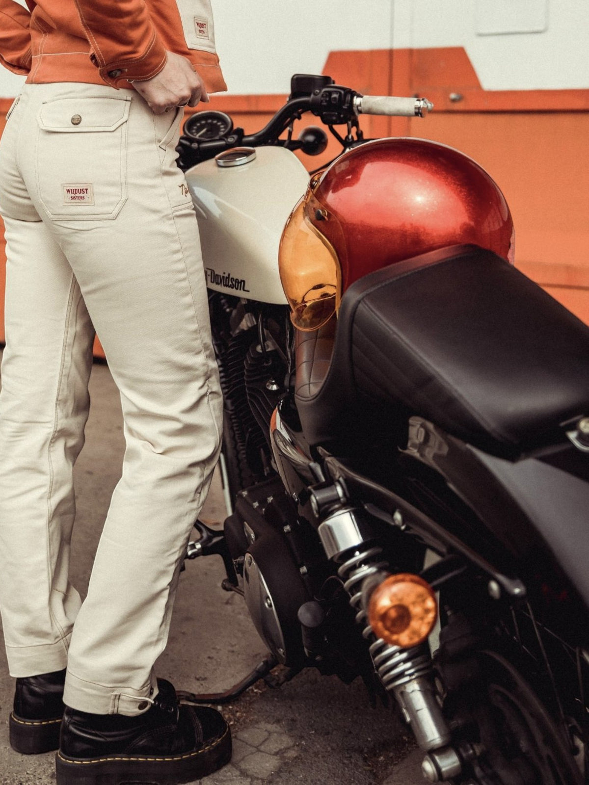 Baffle Haus ivory cargo motorcycle trousers with patch pockets and embroidery, worn by a woman next to a motorcycle.