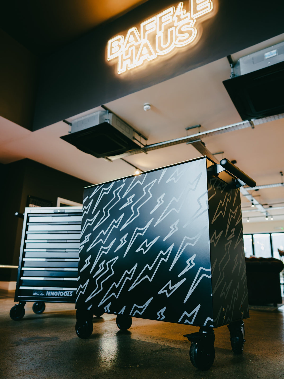 Baffle Haus black metal tool box with white lightning bolt pattern, standing in a workshop with a neon sign above.