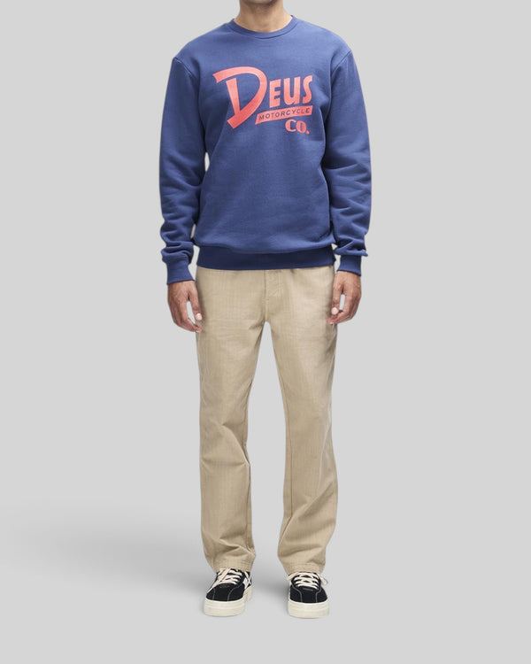 Baffle Haus Trafalgar crew fleece in mechanic blue with Deus graphic print, worn with beige trousers and sneakers