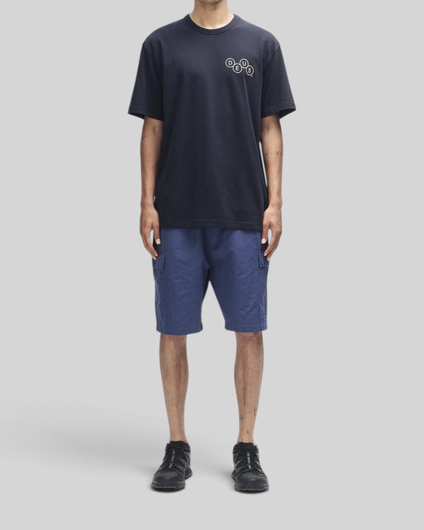 Baffle Haus Mechanism Tee in black organic cotton with chest graphic, worn with blue cargo shorts and black sneakers.