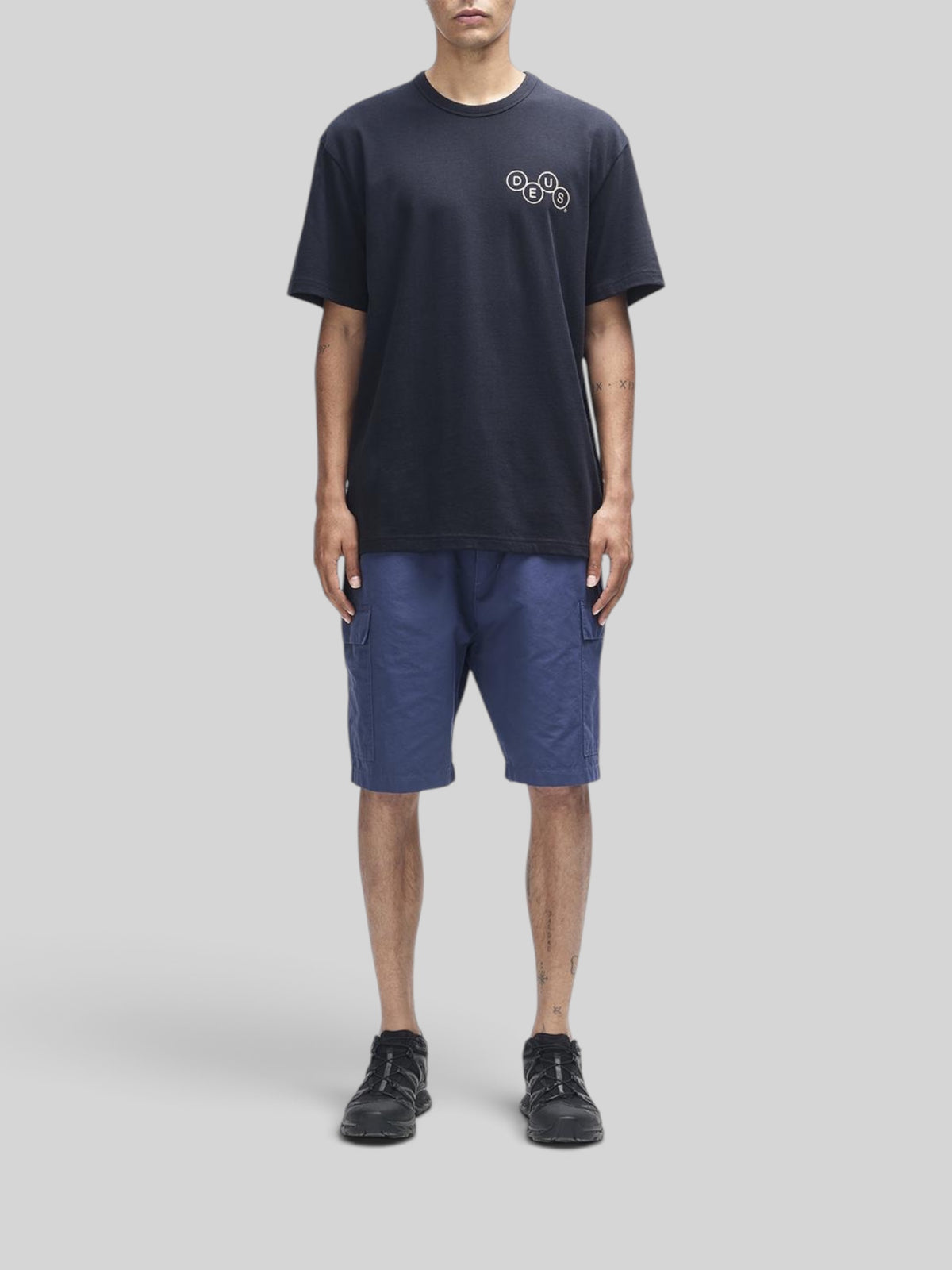 Baffle Haus Mechanism Tee in black organic cotton with chest graphic, worn with blue cargo shorts and black sneakers.