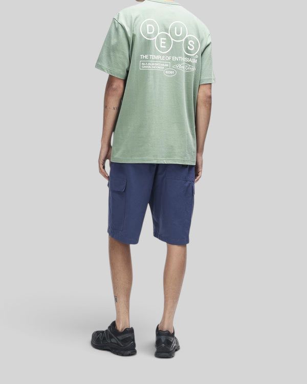 Baffle Haus Mechanism Tee in loden frost green with back graphic print, worn with navy cargo shorts and black sneakers