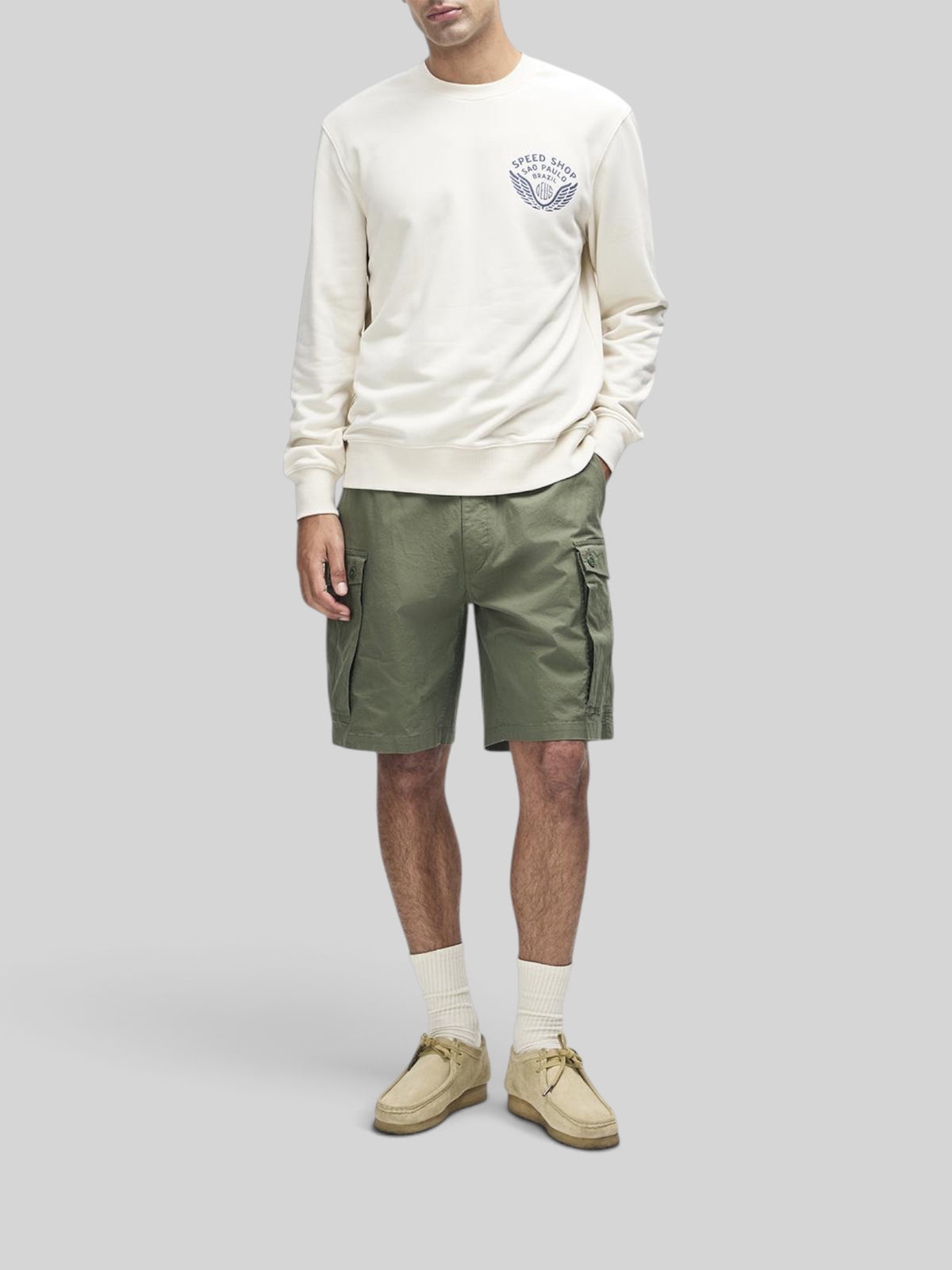 Baffle Haus Munificence crew fleece in dirty white with chest graphic, worn with olive cargo shorts and suede shoes.