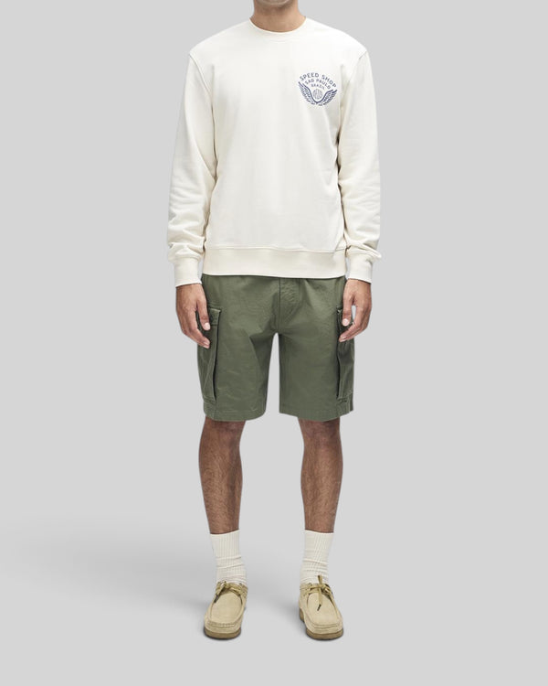 Baffle Haus Munificence Crew dirty white fleece with chest graphic, worn with olive cargo shorts and beige shoes.