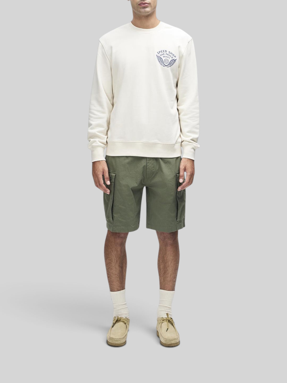 Baffle Haus Munificence Crew dirty white fleece with chest graphic, worn with olive cargo shorts and beige shoes.
