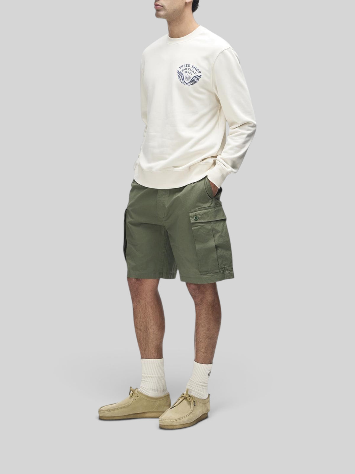 Baffle Haus Munificence crew fleece in dirty white with chest graphic, worn with olive cargo shorts and suede shoes.