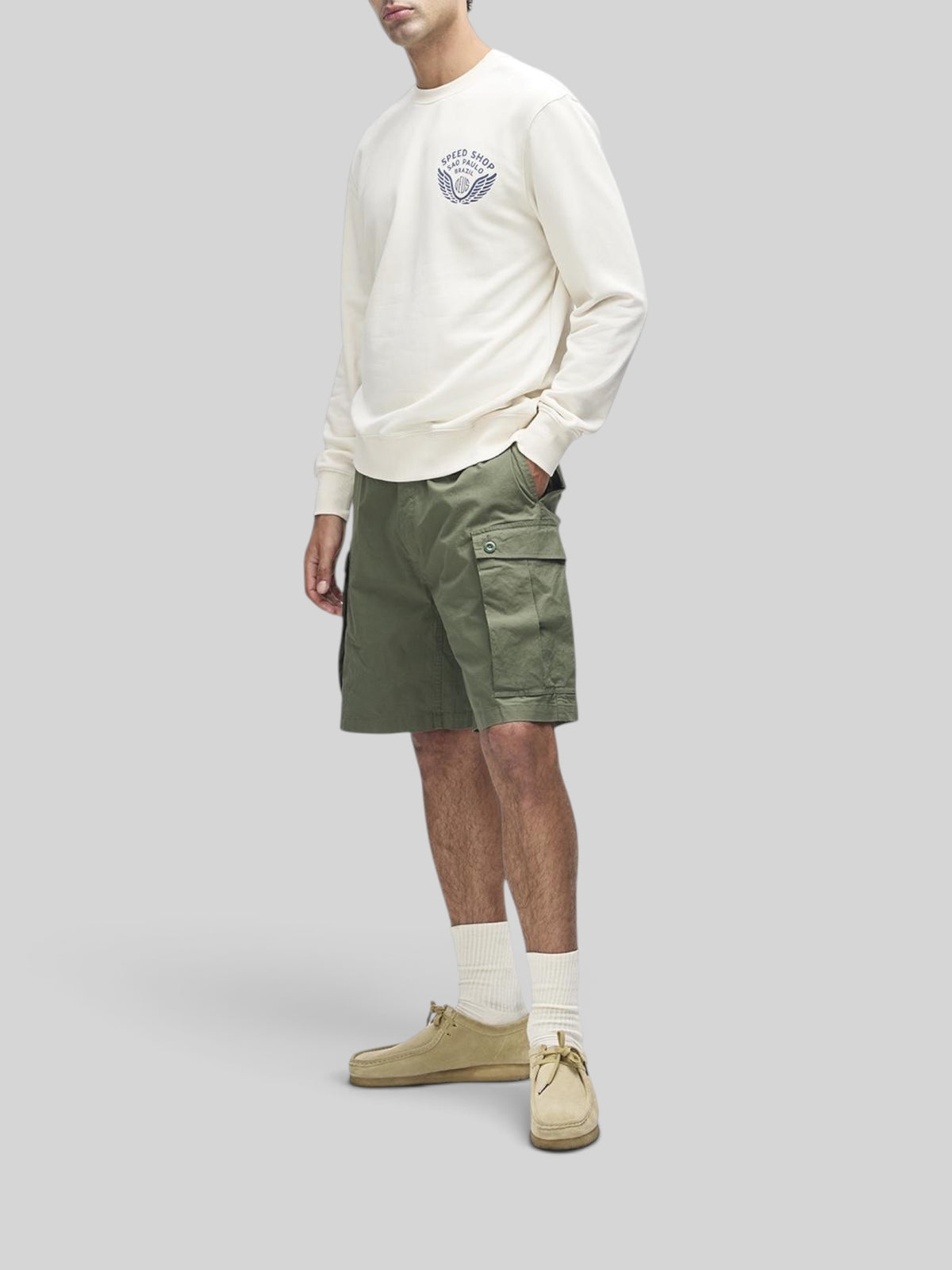 Baffle Haus Munificence crew fleece in dirty white with chest graphic, worn with olive cargo shorts and beige shoes.