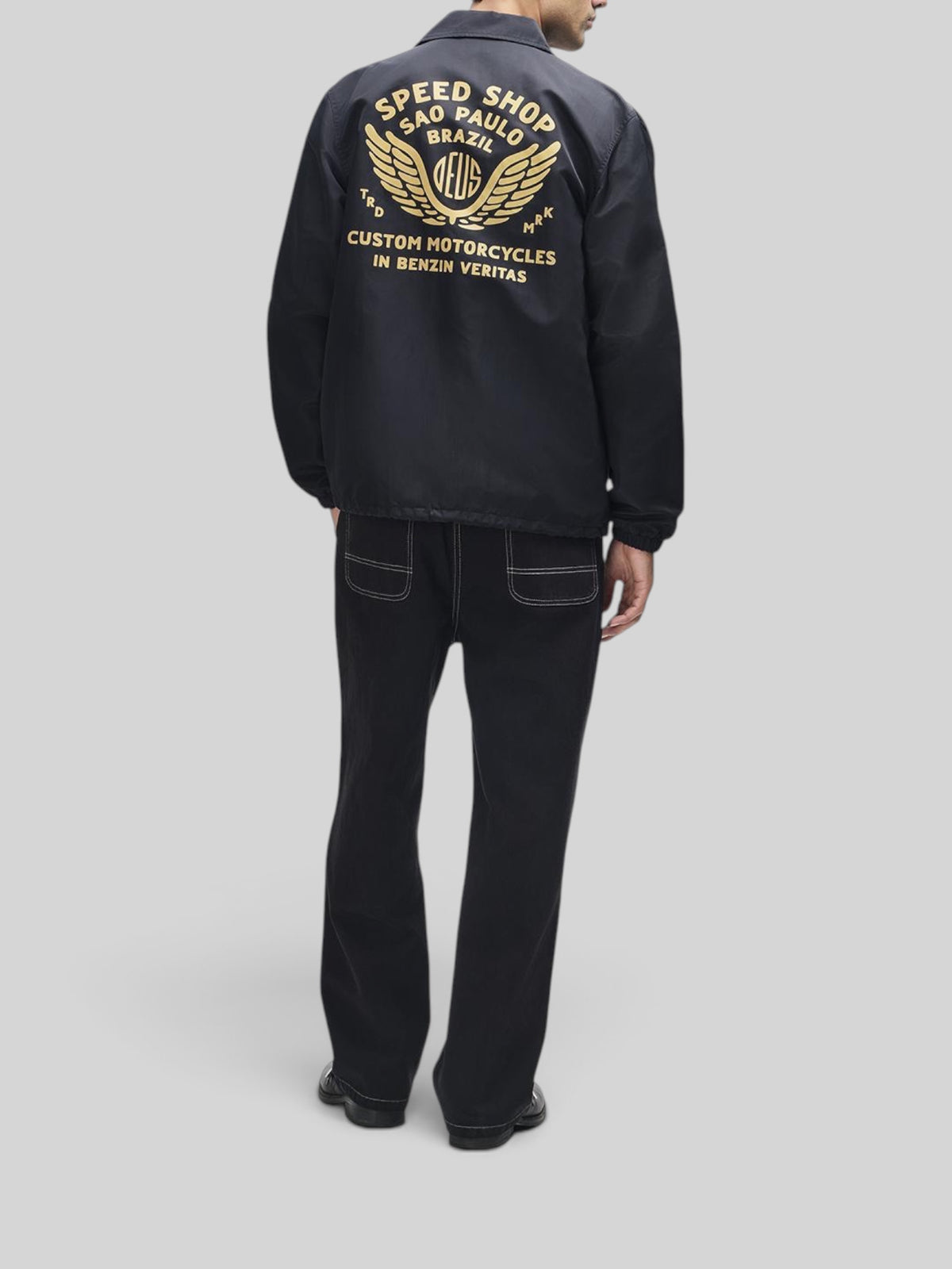Baffle Haus Munificence nylon coach jacket in black with gold graphic print on back, worn by a man from behind.