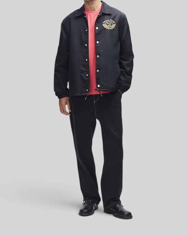 Baffle Haus Munificence nylon coach jacket in black with chest graphic, worn over a red t-shirt and black trousers.