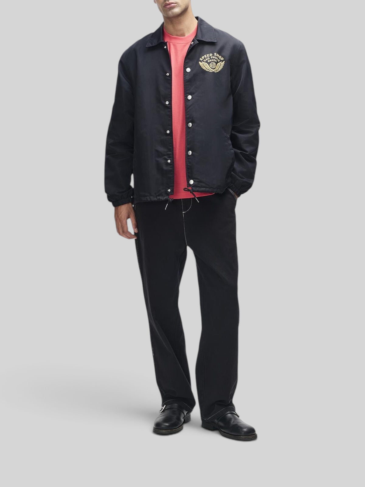 Baffle Haus Munificence nylon coach jacket in black with chest graphic, worn over a red t-shirt and black trousers.