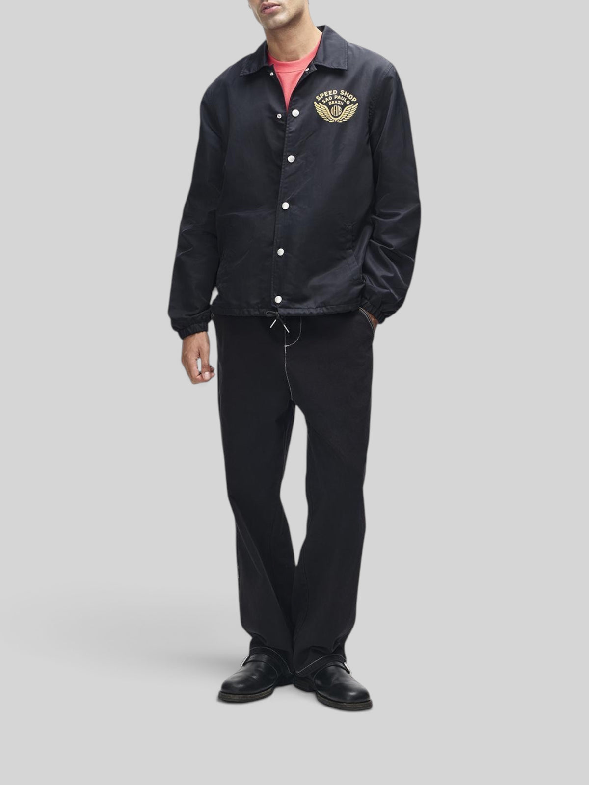Baffle Haus Munificence nylon coach jacket in black with chest graphic, snap buttons, and regular fit on a model.