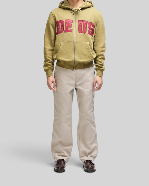 Baffle Haus olive green zip-up hoodie with red chest appliqué, worn by a model with beige trousers.