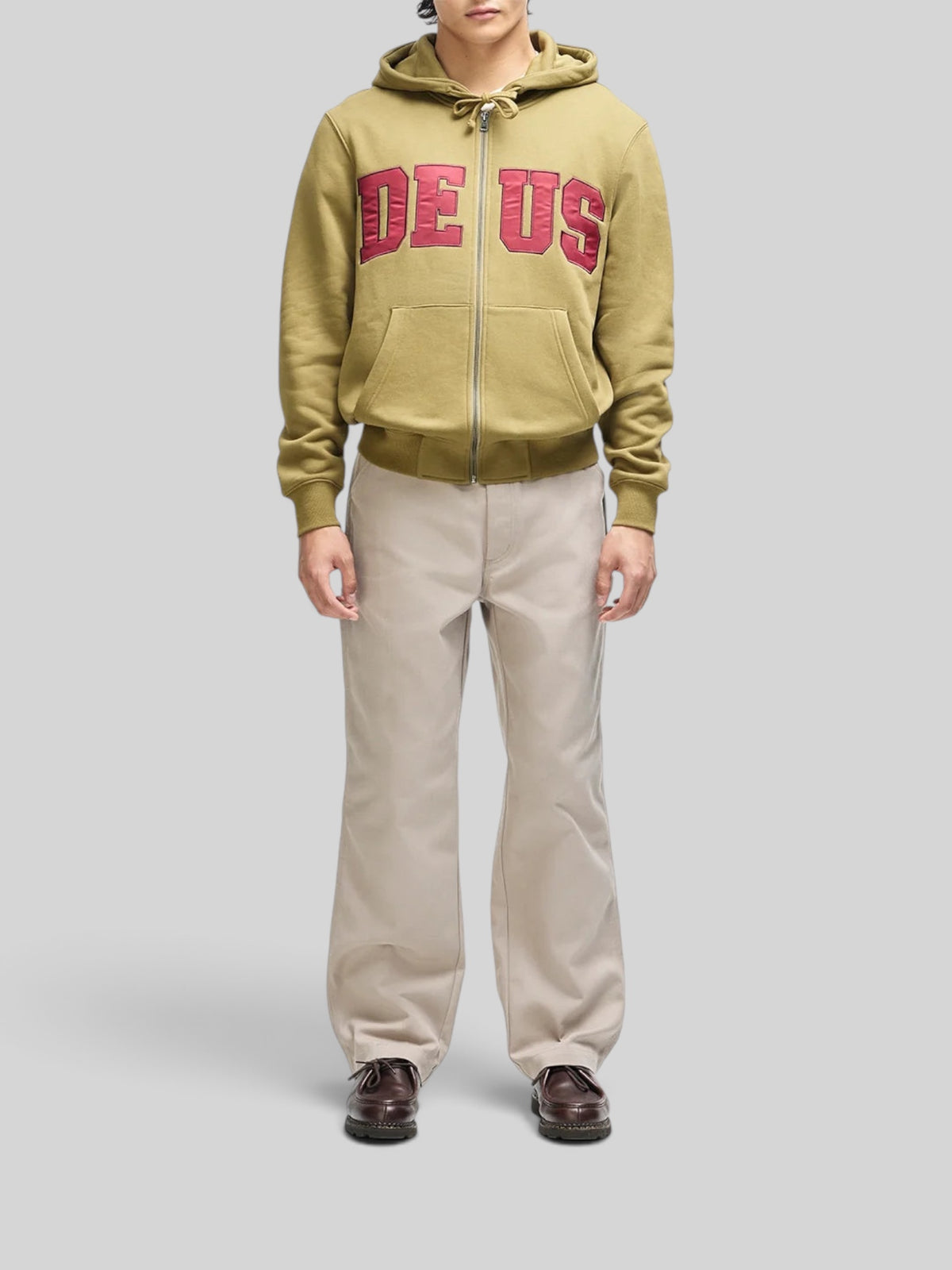 Baffle Haus olive green zip-up hoodie with red chest appliqué, worn by a model with beige trousers.