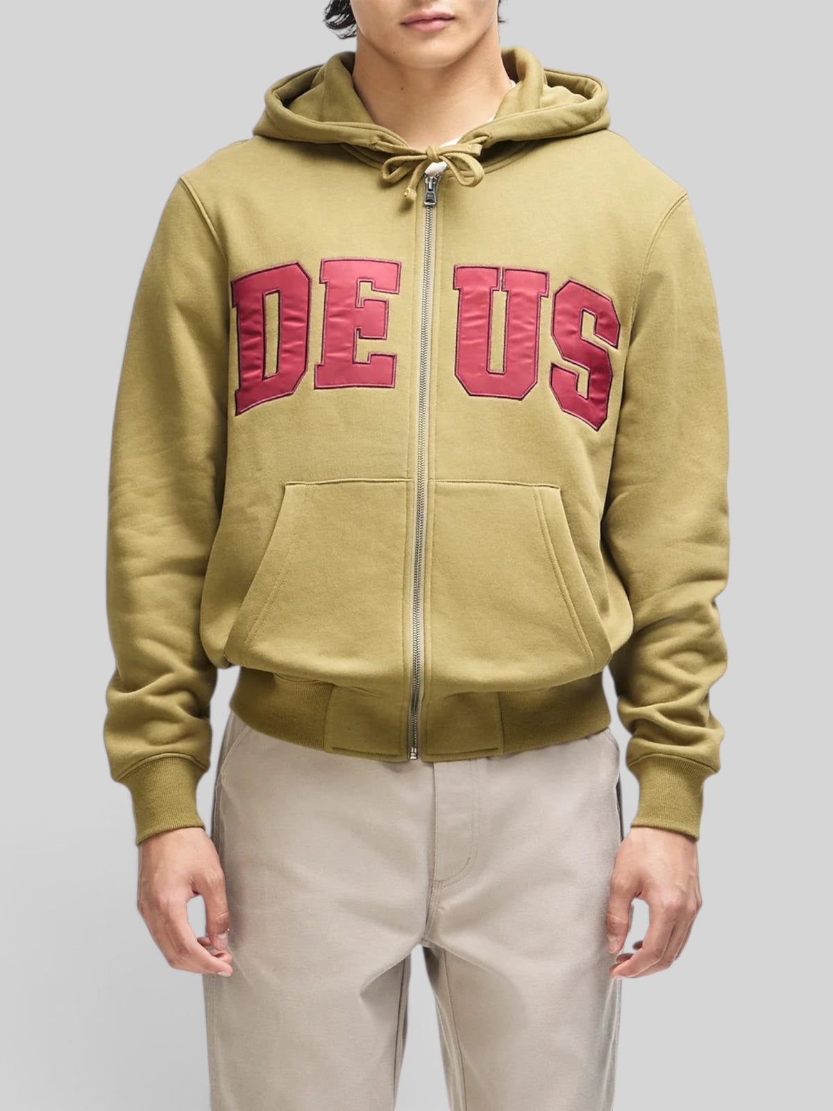 Baffle Haus olive green zip-up hoodie with red DEUS chest appliqué, kangaroo pocket, and ribbed cuffs on a model