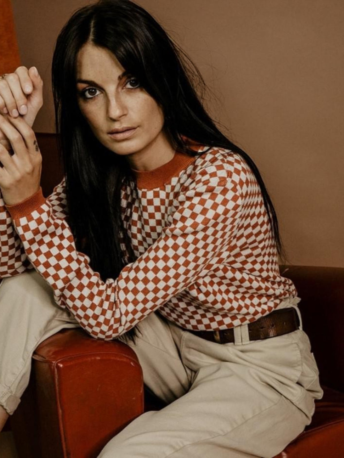 Baffle Haus rust and white chequered knit jumper with ribbed cuffs worn by a woman sitting in a leather chair