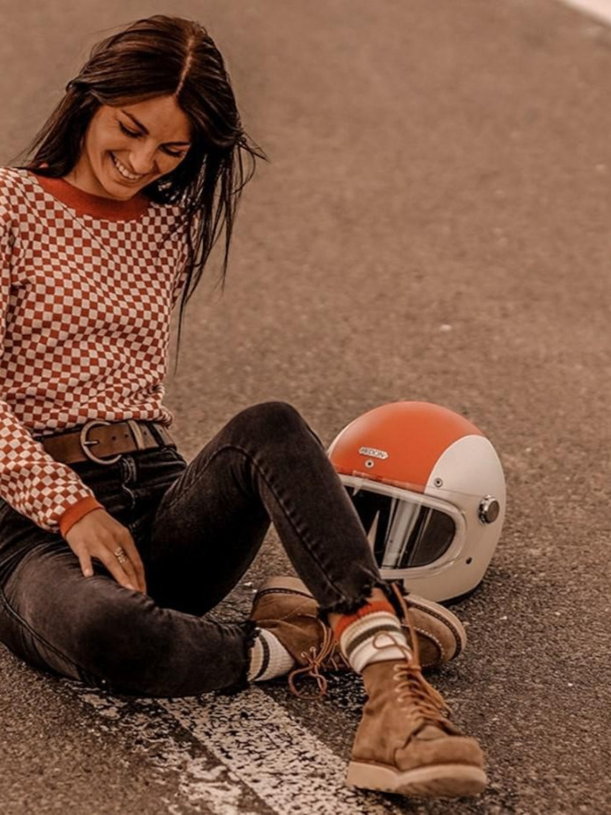 Baffle Haus rust and white chequered racing jumper worn by a woman sitting on a road next to a vintage helmet.