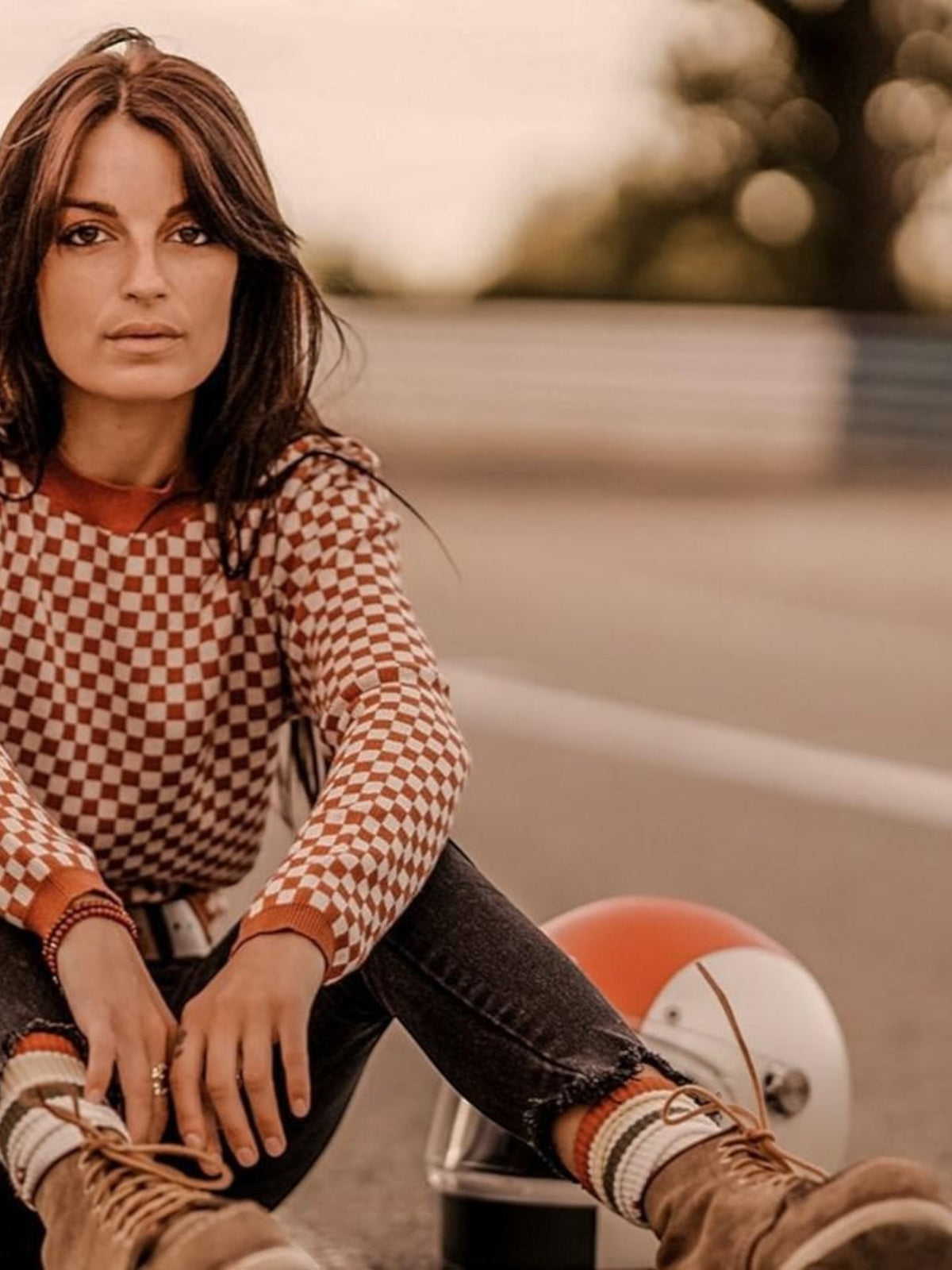 Baffle Haus rust and white chequered racing jumper worn by a woman sitting on a racetrack with a vintage helmet