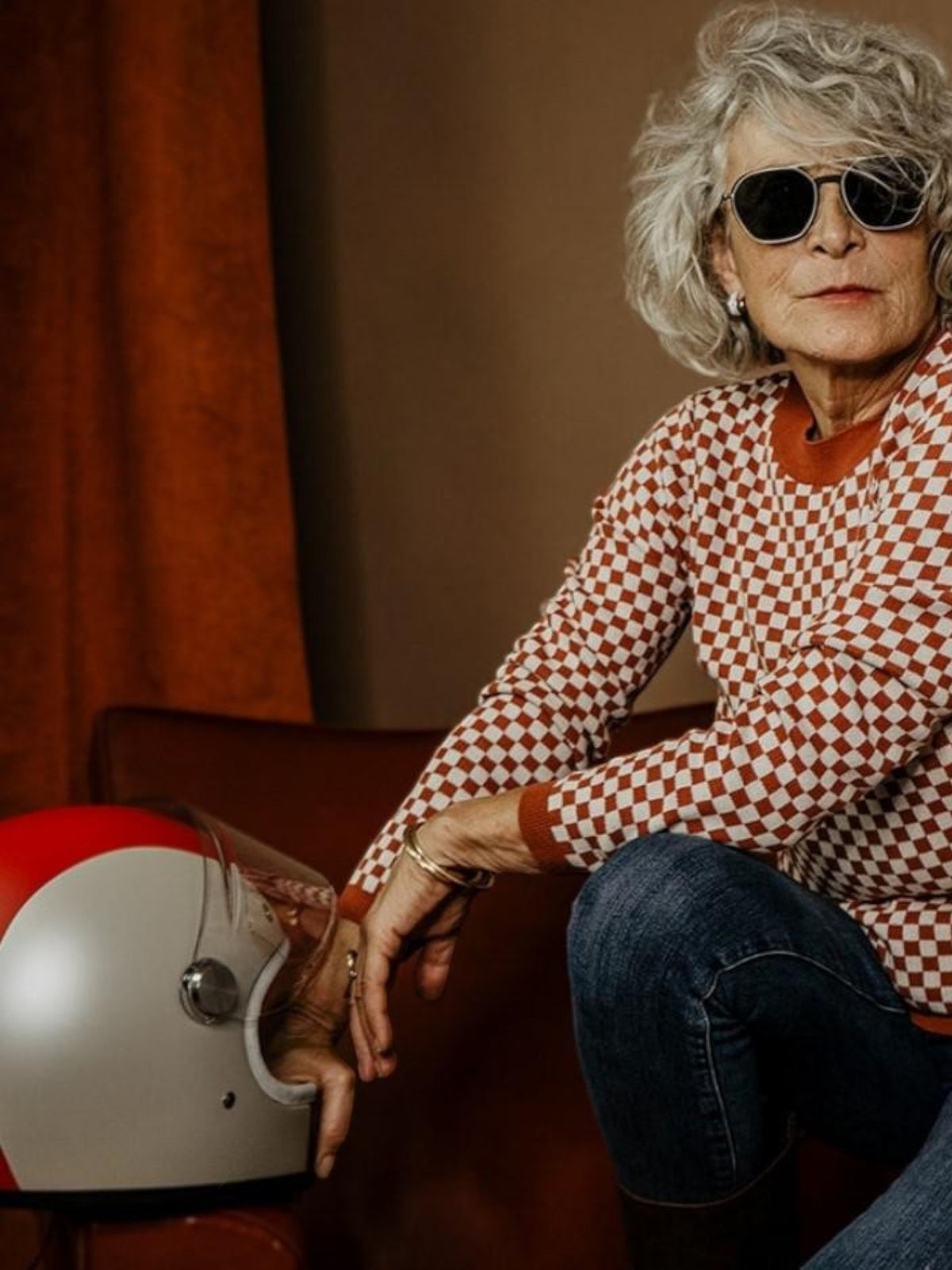 Baffle Haus rust and white chequered racing jumper worn by a woman with sunglasses holding a vintage motorcycle helmet