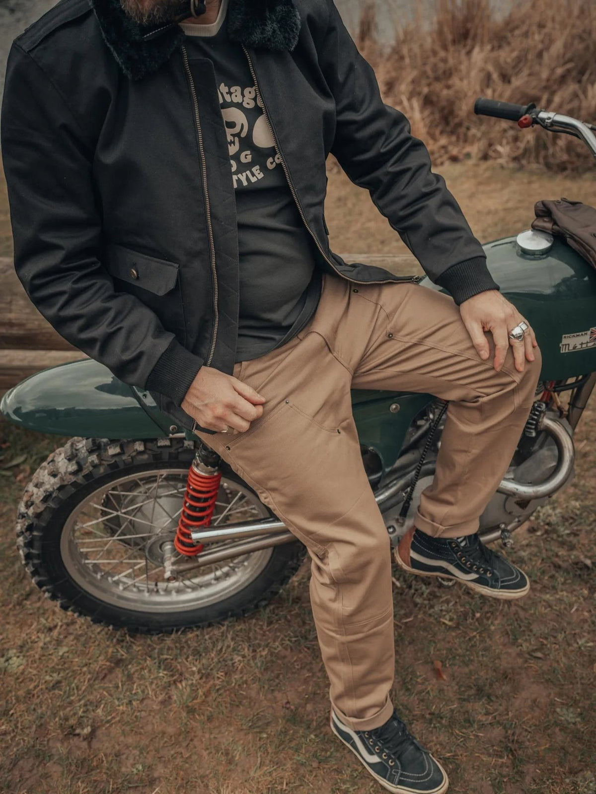 Baffle Haus Service Pants in desert khaki worn by a motorcyclist sitting on a vintage green motorcycle
