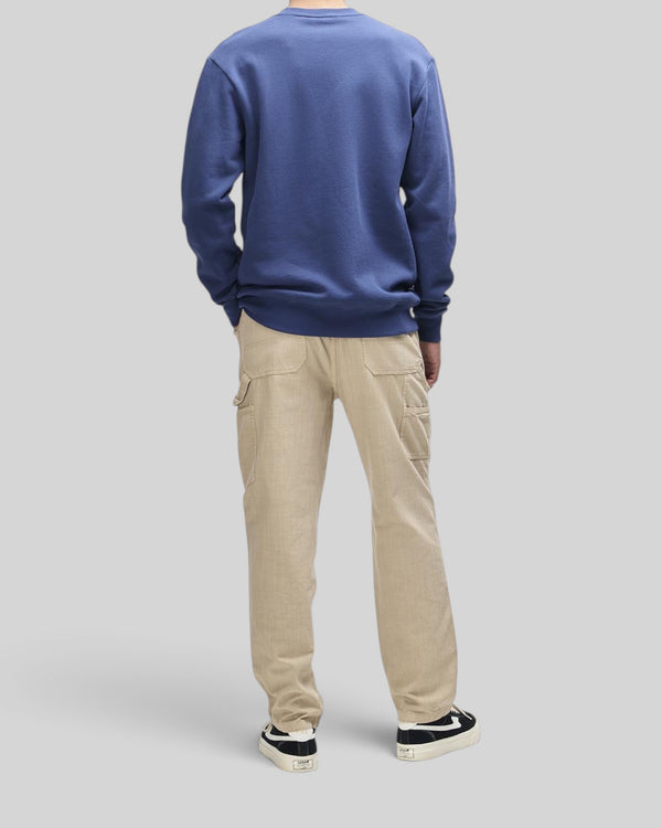 A man wearing a Baffle Haus Trafalgar Crew mechanic blue fleece sweatshirt and beige cargo pants from behind.