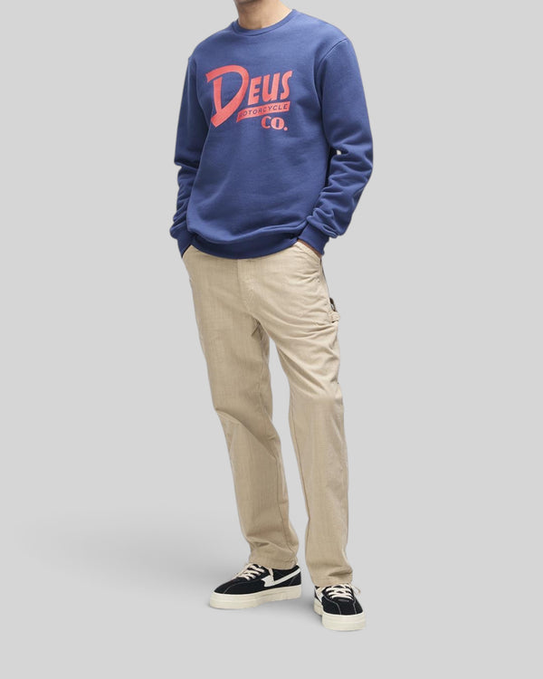 Baffle Haus Trafalgar Crew fleece in mechanic blue with red chest graphic, worn by a model with beige trousers.