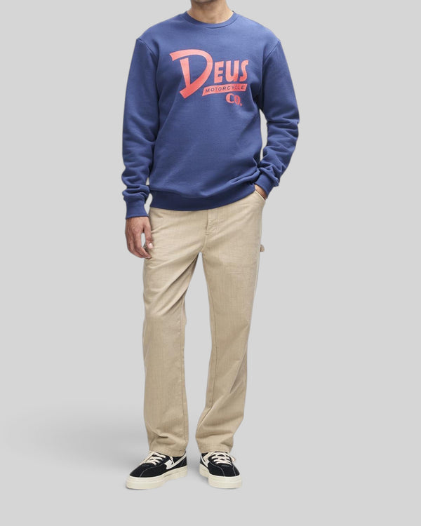 Baffle Haus Trafalgar crew fleece in mechanic blue with chest graphic, worn with beige trousers and sneakers.