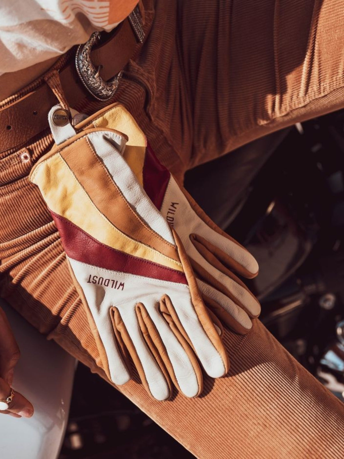 Baffle Haus Wildust 70s stripe leather motorcycle gloves in white, yellow, and red resting on corduroy trousers