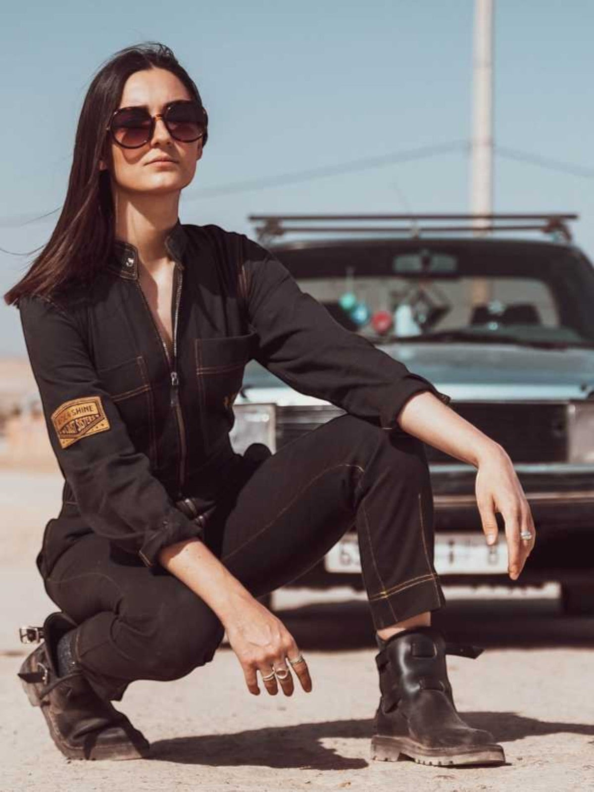 Baffle Haus Wildust black cotton aviator jumpsuit with camel stitching and arm patch, worn by a woman in the desert.