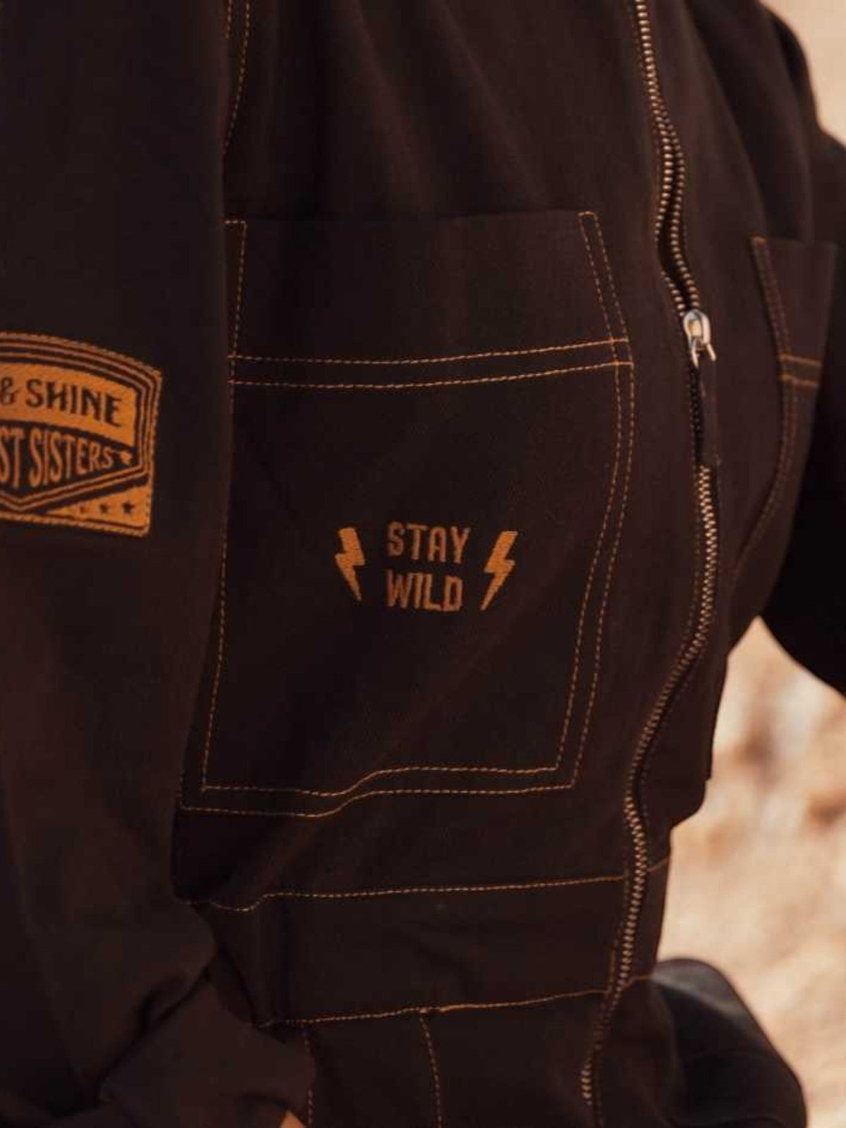 Baffle Haus Wildust black cotton aviator jumpsuit featuring camel stitching, chest embroidery, and arm patch detail.